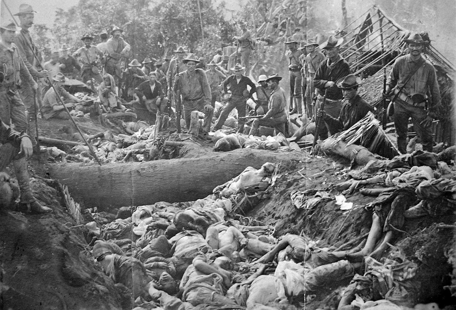Black and white photo of American soldiers standing above hundreds of bodies of dead Filipinos